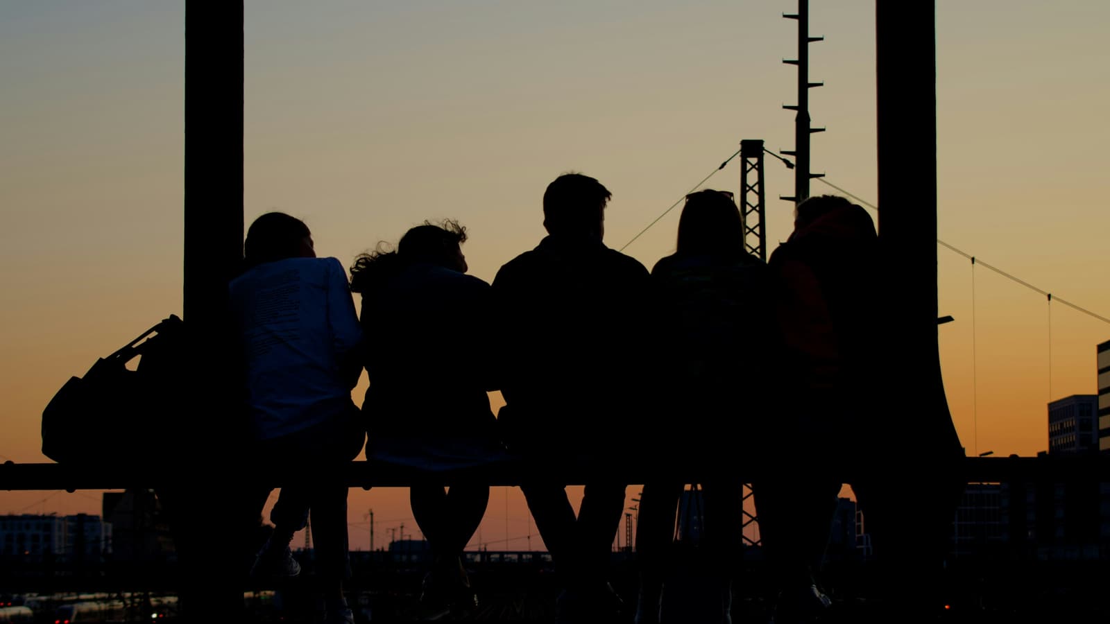 Five friends sit side by side on a bridge railing, silhouetted against an ember-orange sunset