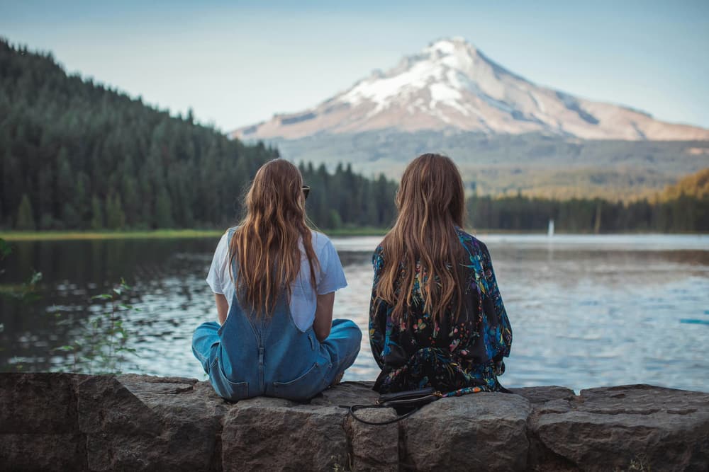 Two friends sit on a stone wall beside a still lake, looking out at a snow-capped mountain