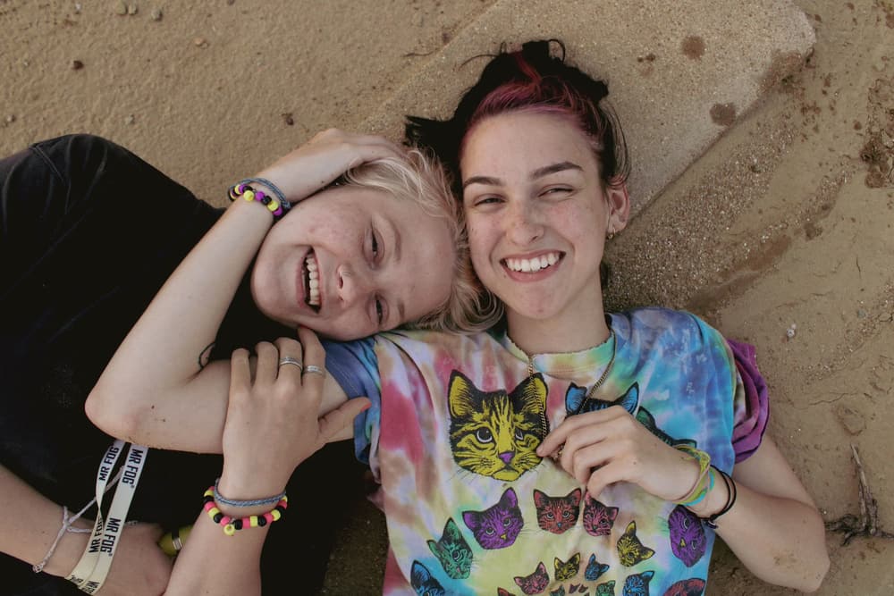 Two friends lying on warm sand, heads together, laughing up at the camera